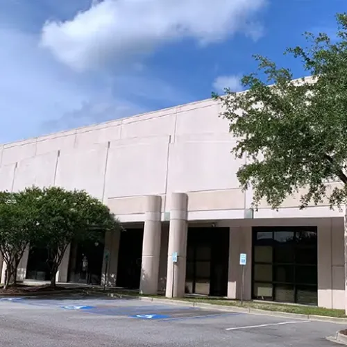 A lightcolored building with pillars stands under trees on a paved street A blue handicapped parking sign is visible