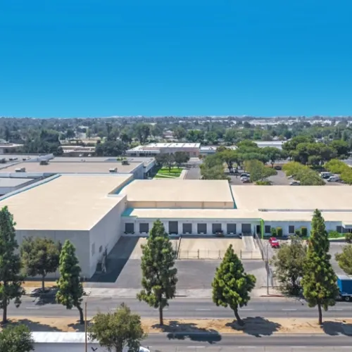 A long white warehouse complex sits under a bright sky with tall trees lining the road in Fresno California