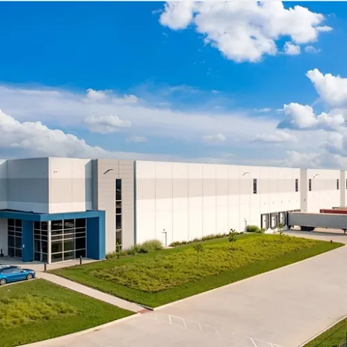 White industrial buildings line a paved road with a grassy median A large blue truck sits near one of the buildings