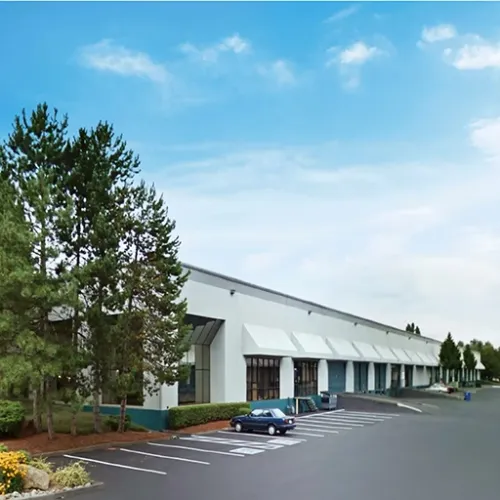 A large white commercial building with rollup doors sits amongst mature evergreen trees and landscaping in Kent Washington