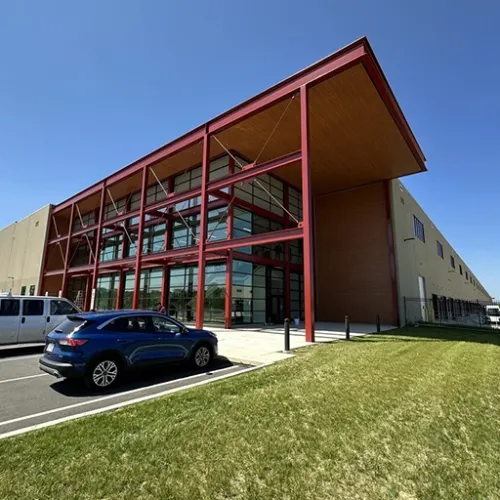 A modern large warehouse building with a light gray facade and prominent red steel supports sits on a grassy lawn A white