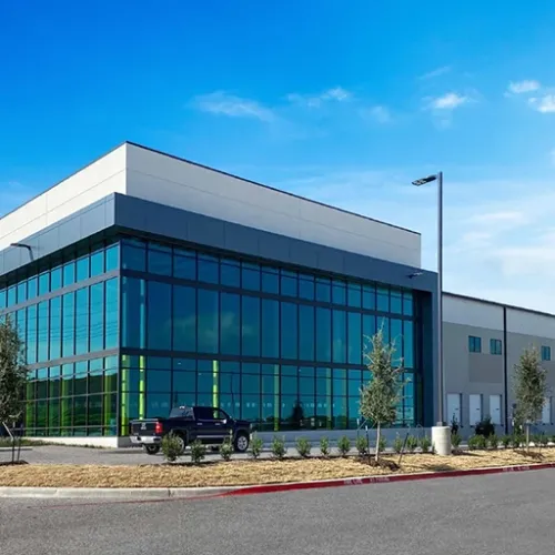 A modern lightcolored building with extensive glass panels sits on a paved lot Landscaping and a street are visible in the