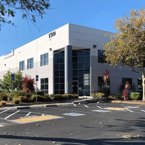A modern lightcolored commercial building with large windows sits on a paved lot surrounded by shrubs and red plants