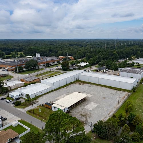 Aerial view of industrial complex with large white L-shaped warehouse, concrete lot, and smaller buildings surrounded by trees.