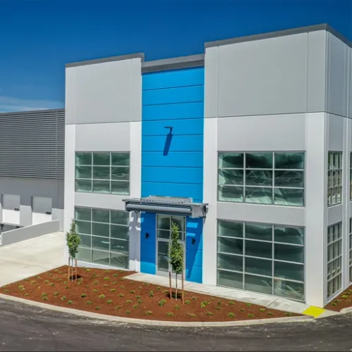 A modern blueandwhite commercial building with a small entranceway sits on an asphalt lot dotted with red ground cover and