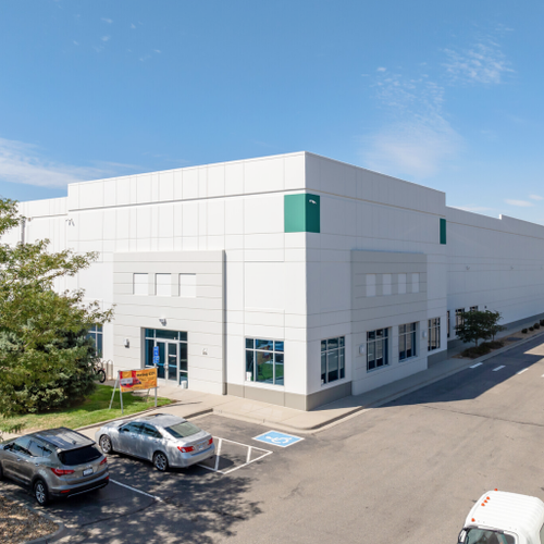 A large white commercial building with green accents sits on an asphalt road a silver car is parked nearby