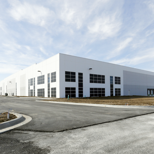 A large modern white warehouse stands prominently on an asphalt road with a grassy median Numerous windows punctuate the