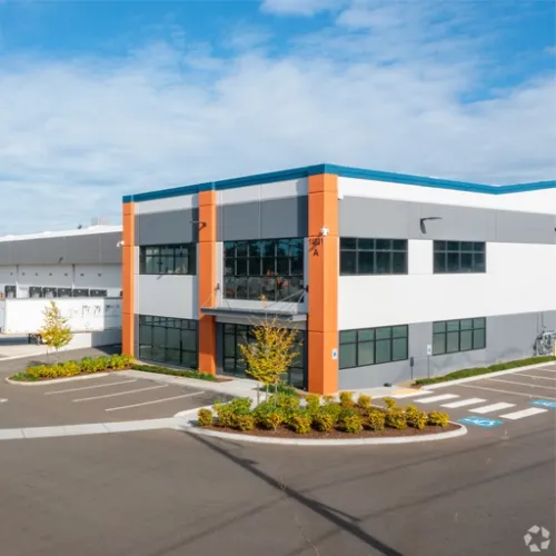 A modern lightcolored building with orange accents sits on an asphalt parking lot surrounded by landscaping
