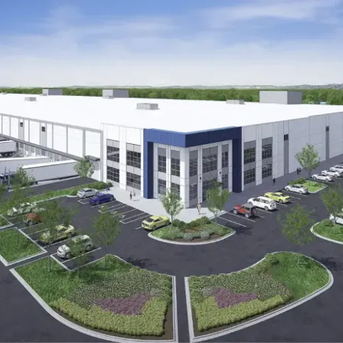 A modern warehouse building with a blue roof stands prominently against a green landscape featuring parking areas and