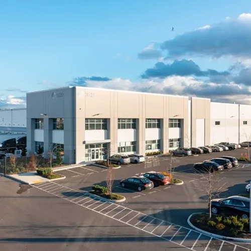 A large modern warehouse building with expansive parking lot stripes and several parked cars dominates the scene